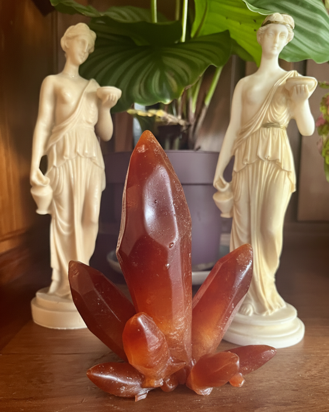 Modèle de krystal, adaptation Cornaline. Objet en silicone, souple d'un objet intime en forme de de krystal posé sur une bibliothèque en bois, s'intégrant dans le décor. Dans l'arrière plan sont deux statuettes grecque et une plant verte, flouté afin de garder le focus sur le krystal de la marque Playsir