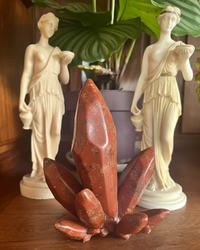 Modèle de krystal, adaptation Jaspe rouge. Objet en silicone, souple d'un objet intime en forme de de krystal posé sur une bibliothèque en bois, s'intégrant dans le décor. Dans l'arrière plan sont deux statuettes grecque et une plant verte, flouté afin de garder le focus sur le krystal de la marque Playsir