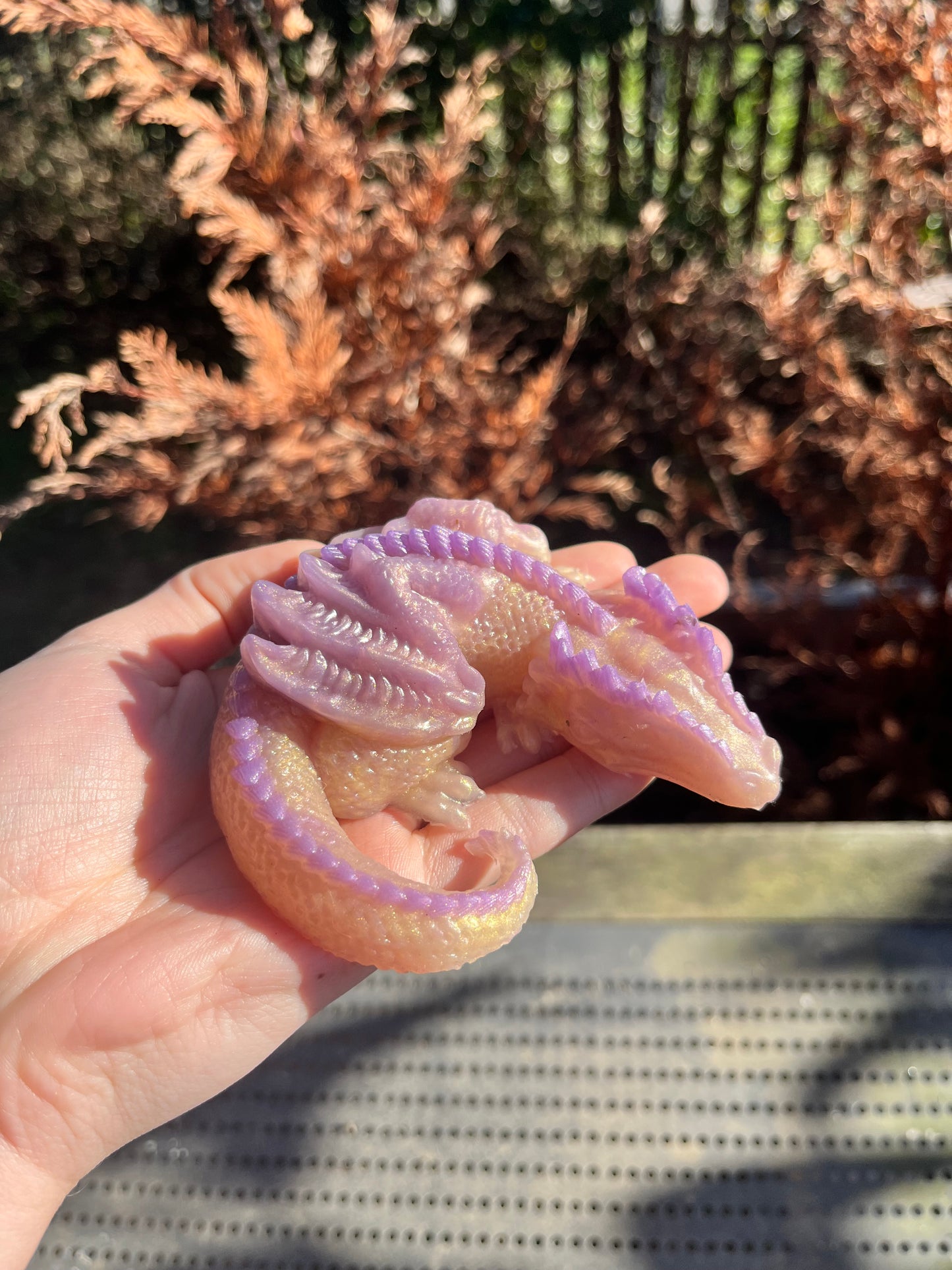 jouet anti stresse de la marque Playsir. En silicone coloré en or et violet irisé. Jouet Dragon posé sur une main pour comprendre la taille, en arrière plan il y a un décor naturel extérieur.