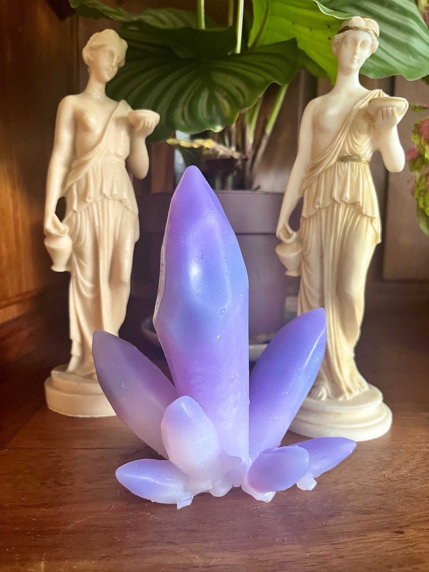 Photo de l'objet krystal de la marque Playsir, un objet intime décoratif fabriqué à la main avec du silicone certifié pour un usage sécurisé intime. krystal s'inspire de la lithothérapie et prend ici la forme d'un améthyste. Il est mis en scène sur une bibliothèque en bois massif devant deux statuettes grecques et une plante en pot.
