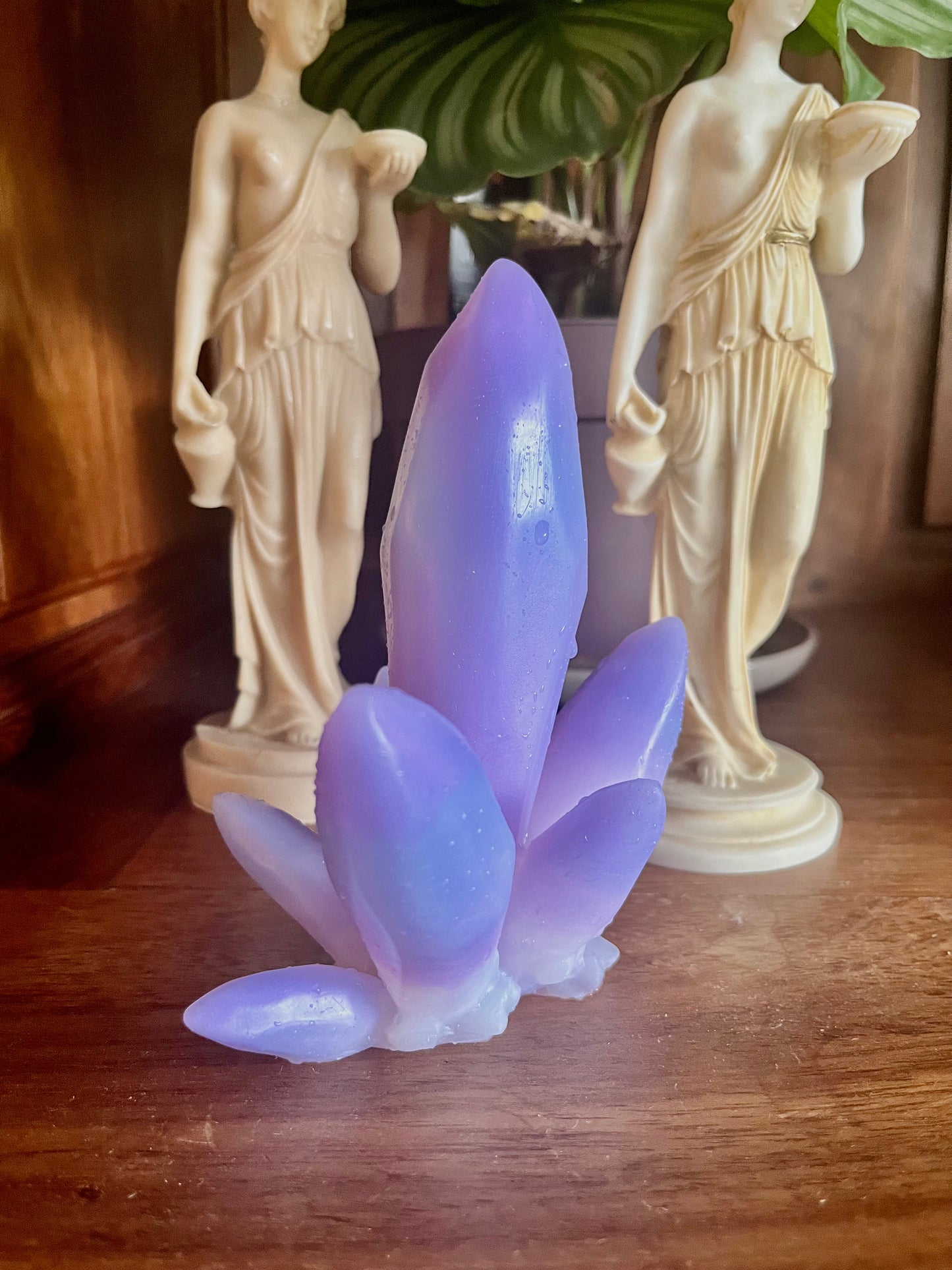 Objets Krystal vue de coté , aux inspiration et apparence d'une pierre précieuse Améthyste. Objet intime décoratif en silicone violet devant un décor de statues grecques et une plate en pot. Le krystal en silicone est posé sur une bibliothèque en bois.