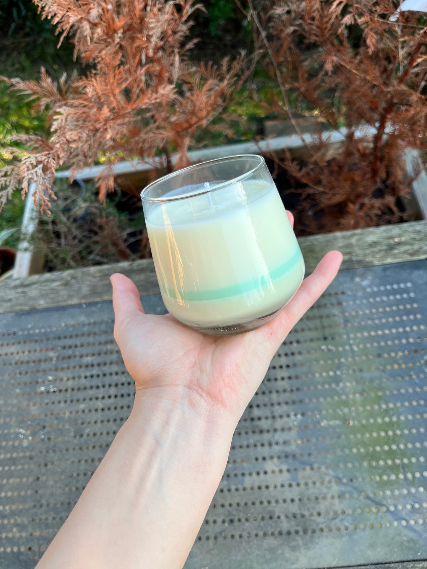 Bougie Malachite, dans un décor extérieur, posé sur une main pour voir le rapport de taille. La photo est lumineuse et le décor naturelle à l'arrière plant est flouter afin de rapporter l'attention sur la bougie de la marque Playsir.